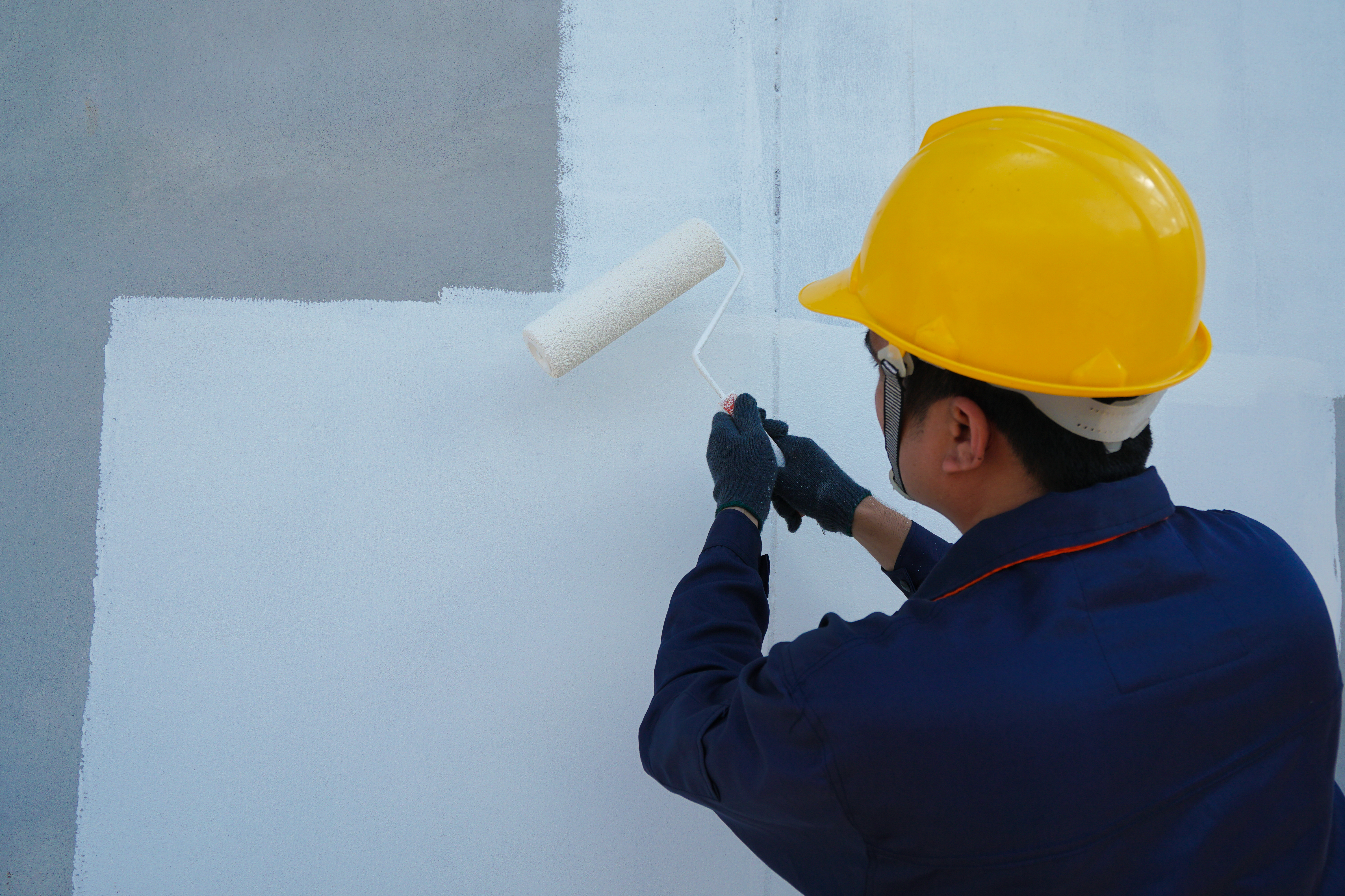 Asian male construction worker is painting with paint roller on white house wall, home decoration and construction concept in project.